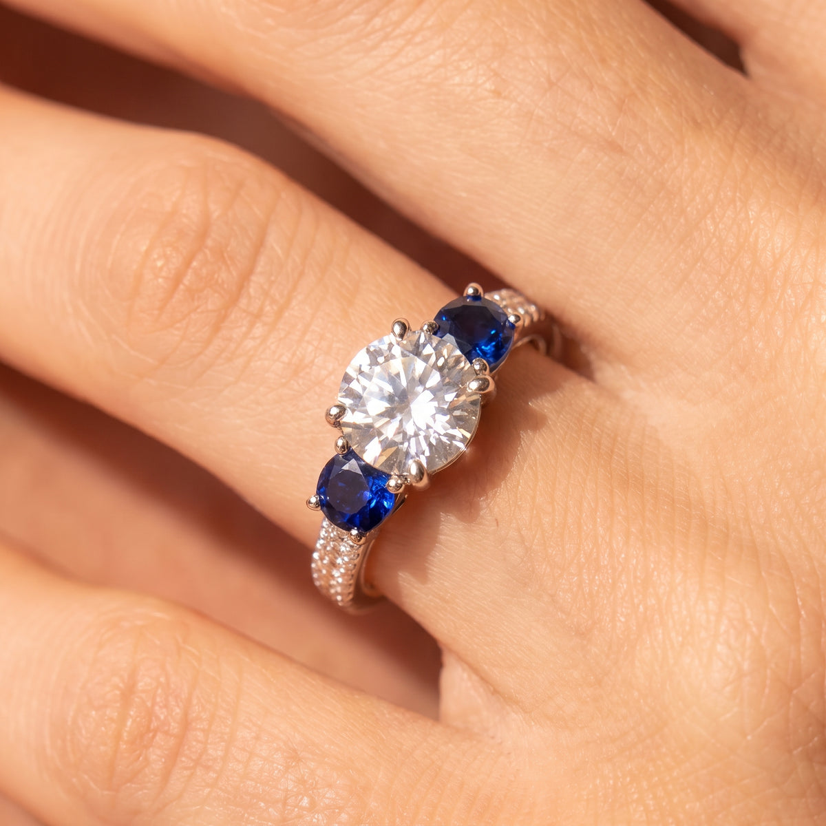 A close-up of a silver-toned ring with a large round clear center stone and two deep blue oval side stones, worn on a hand.
