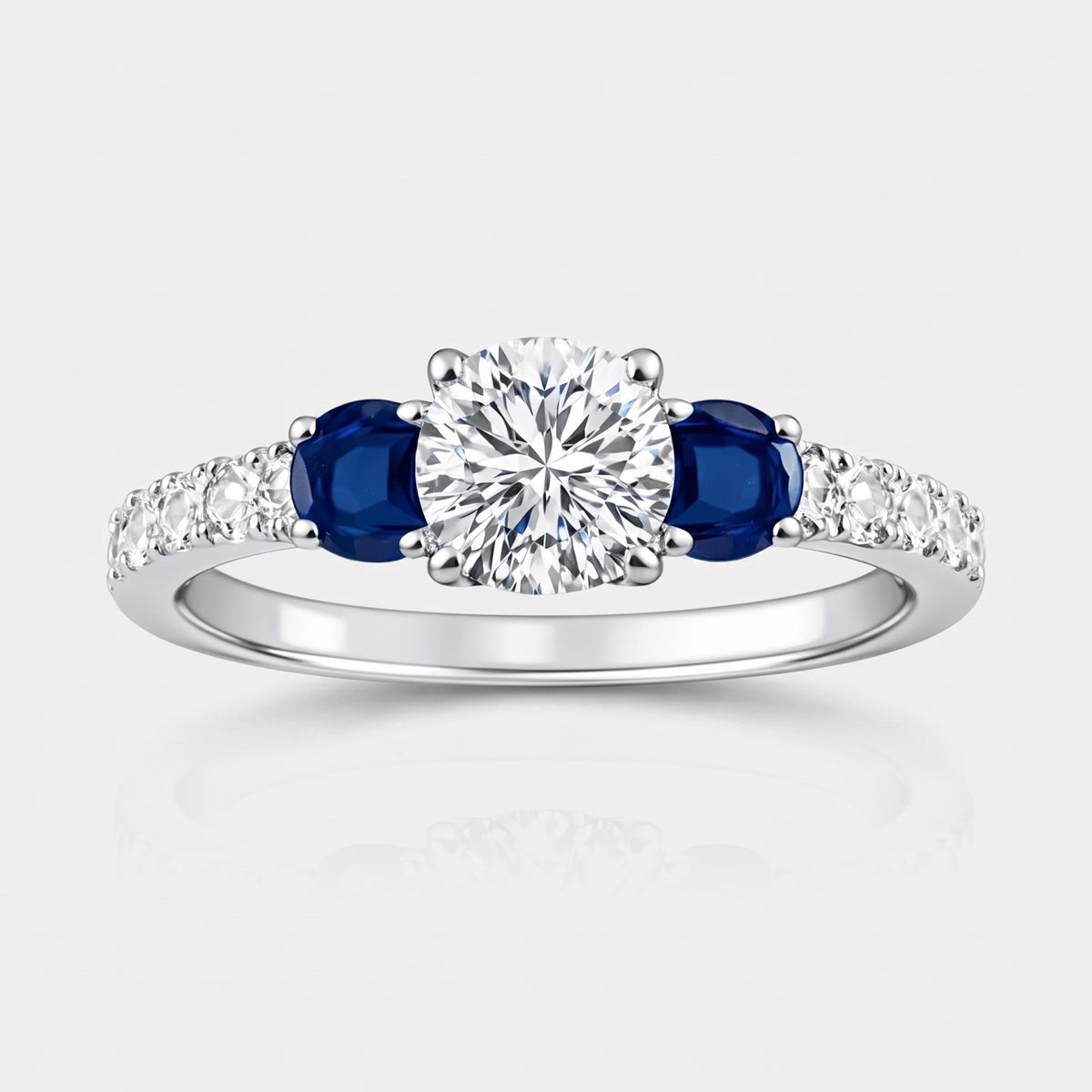 A silver-colored diamond ring featuring a round-cut center diamond flanked by two oval-cut deep blue sapphires, with small pavé diamonds along the shoulders, set against a light gray background.
