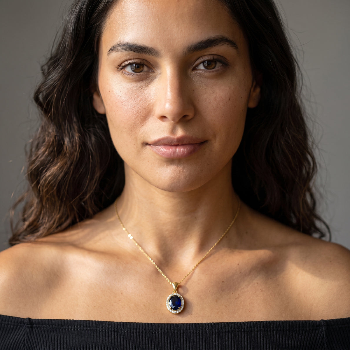A close-up portrait of a woman wearing a delicate gold chain necklace with a teardrop-shaped dark blue gemstone pendant, framed with small clear stones, set against a neutral gray background.