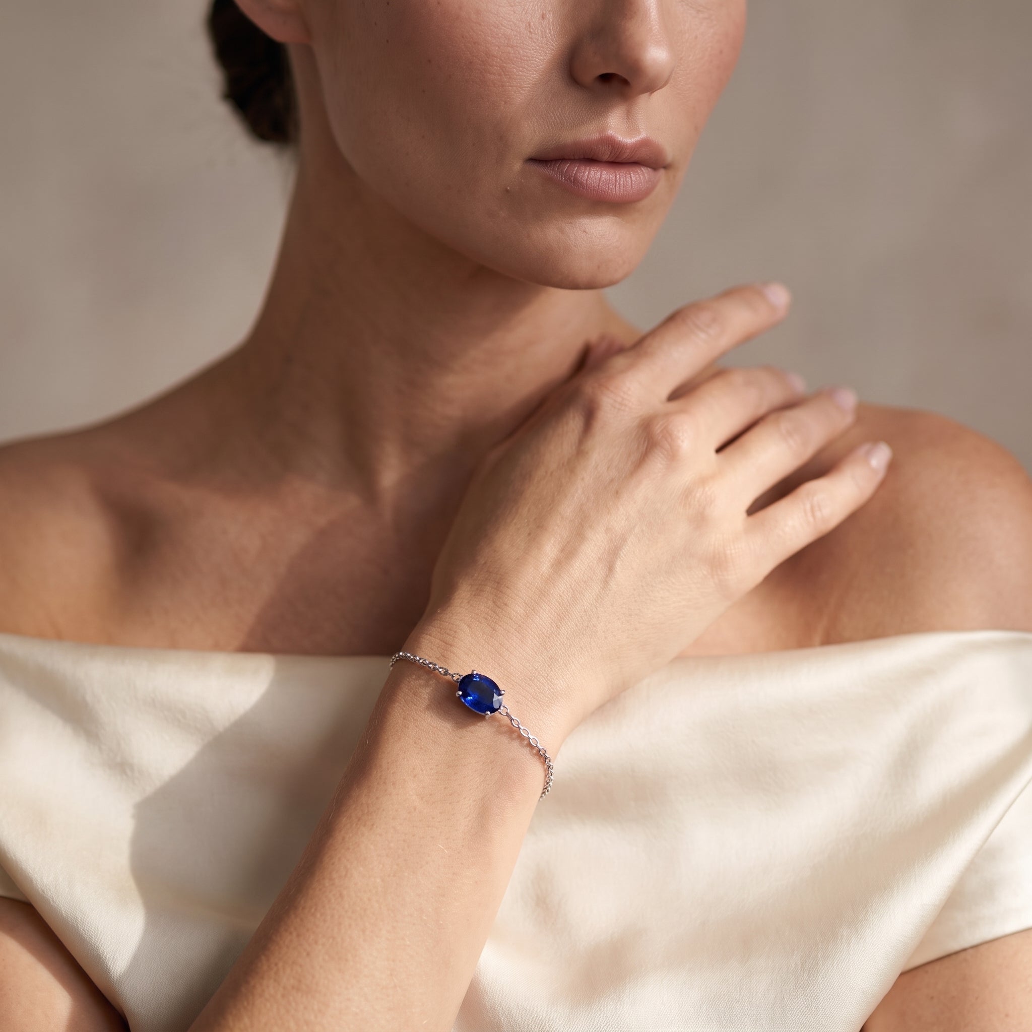 A close-up lifestyle shot of a model wearing a delicate silver chain bracelet with a round, deep blue gemstone centerpiece.