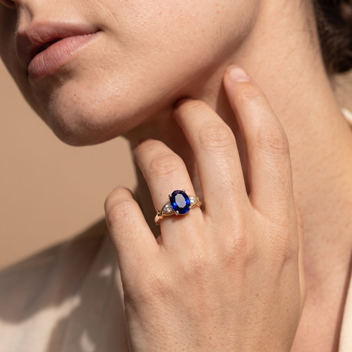 A close-up lifestyle shot of a model wearing a gold ring with a deep blue oval center stone and small side stones, set against a warm beige background.