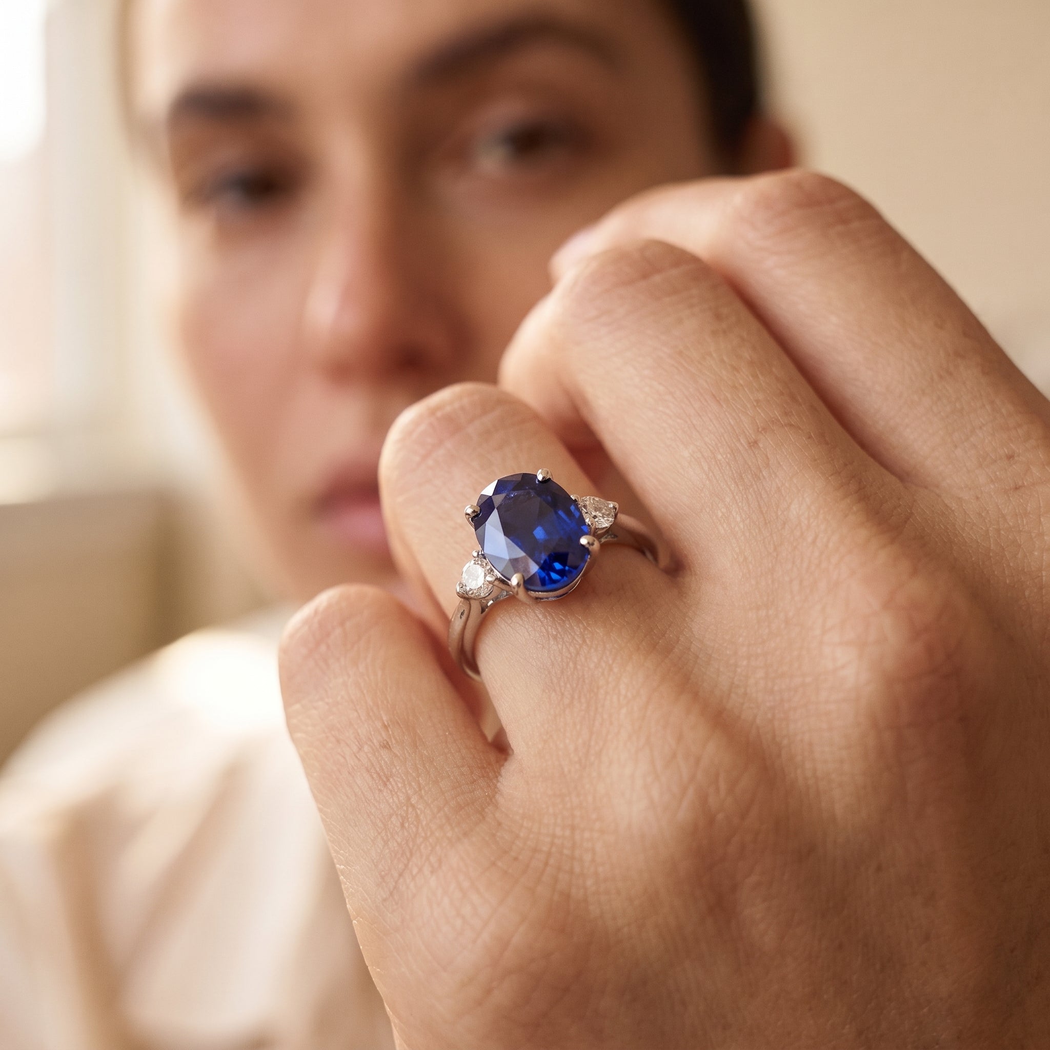 A close-up lifestyle shot of a person wearing a silver ring with a large oval blue sapphire and small side diamonds, with their hand in sharp focus and their face softly blurred in the background.