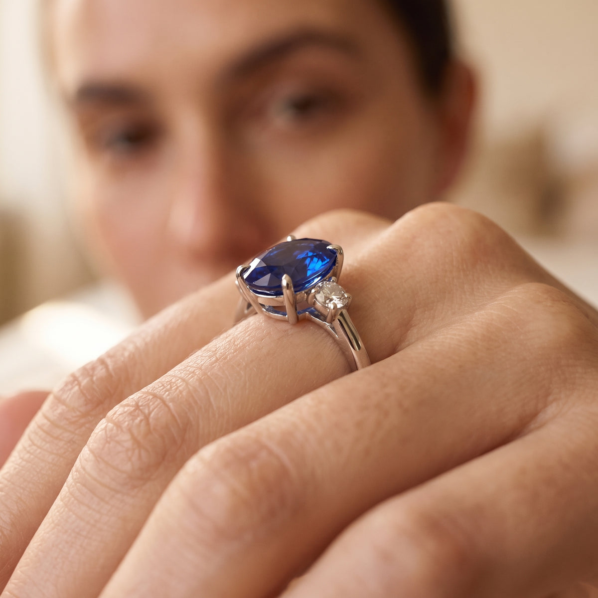 A close-up of a hand wearing a silver ring with a large oval-cut blue gemstone and a smaller clear accent stone, with a softly blurred face in the background.
